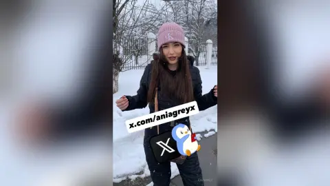 Young woman in a black winter jacket and pink beanie smiling in a snowy outdoor park setting.