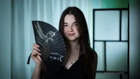 Young woman in a black dress holding a decorative folding fan in a soft, moody indoor setting.