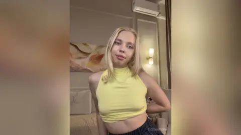 Blonde woman in a yellow halter crop top posing in a bright, modern bedroom.
