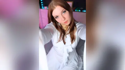 Young woman with long brown hair wearing a white ruffled blouse in a soft pink lit room.