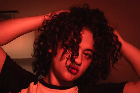 Young woman with curly hair in a black t-shirt posing under moody red lighting.