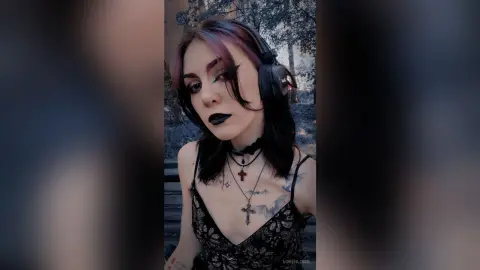 LorenLoiss poses with black lipstick and headphones, wearing a black lace camisole and cross necklaces against a forest background.