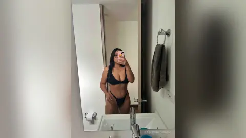 Curvy woman in a black bikini taking a mirror selfie in a modern white bathroom.