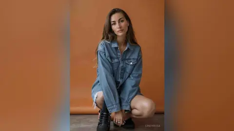 Young woman in a denim jacket and black boots posing against an orange studio background.