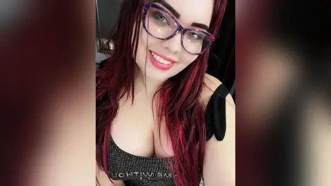 Smiling woman with red hair and purple glasses wearing a black sequin top in a bright indoor setting.