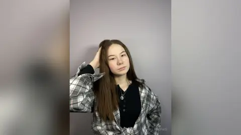 Young woman with long brown hair wearing a black top and grey plaid shirt in a minimalist indoor setting.