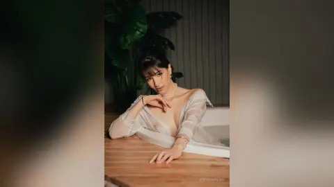 JohanaPiero — Elegant woman in a white sheer blouse leaning on a wooden table in a moody, tropical indoor setting.