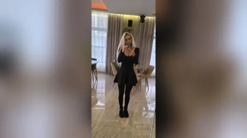 Blonde woman in a chic black long-sleeve mini dress taking a mirror selfie in a bright modern dining room.