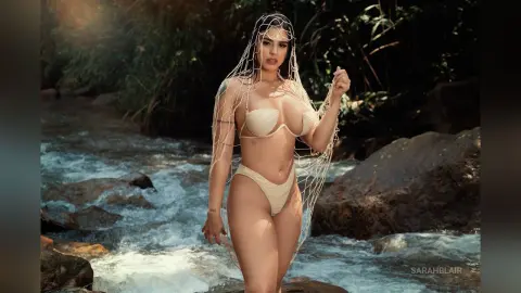 Glamorous woman in a white bikini and sheer mesh cover-up posing by a flowing forest river.