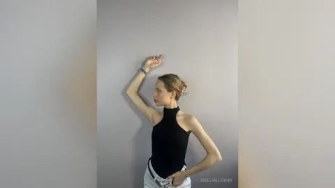MalliaLucioni poses against a grey wall, wearing a black halter neck top and white pants with her arm raised above her head.