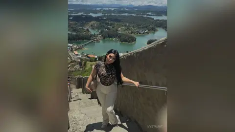 IvyStuart poses outdoors on concrete stairs wearing a leopard print top and white pants, overlooking a scenic lake and green hills.