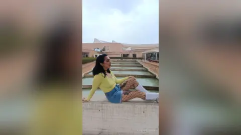 Woman in a yellow long-sleeve top and denim skirt sitting by a modern outdoor water feature under a cloudy sky.