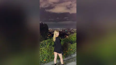 Blonde woman in a chic black mini dress standing against a moody night cityscape view.