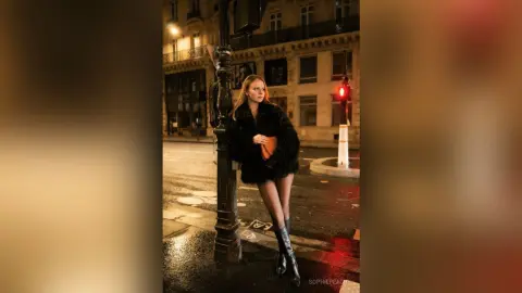 SophiePeachy poses on a city street at night wearing a black fuzzy coat and tall black boots.