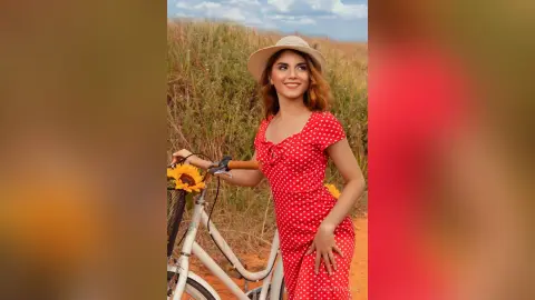ValeryRouge stands outdoors in a red polka-dot dress and a tan sun hat, posing next to a white bicycle in a field.