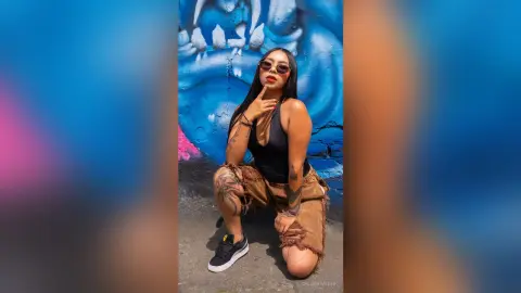 ChloeRiveiro — Stylish woman in black bodysuit and denim shorts posing confidently against a vibrant blue graffiti wall.