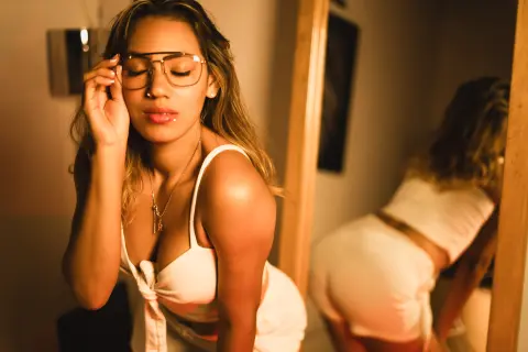 Glamorous woman in white summer outfit and gold glasses posing in warm, moody indoor lighting.