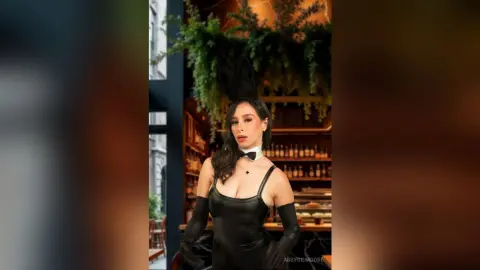 Elegant woman in a black bodysuit and bunny ears posing in a moody, plant-filled lounge setting.