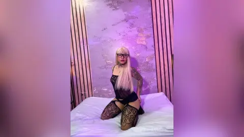 Blonde woman in black lace lingerie and stockings posing on a bed in a moody purple lit bedroom.