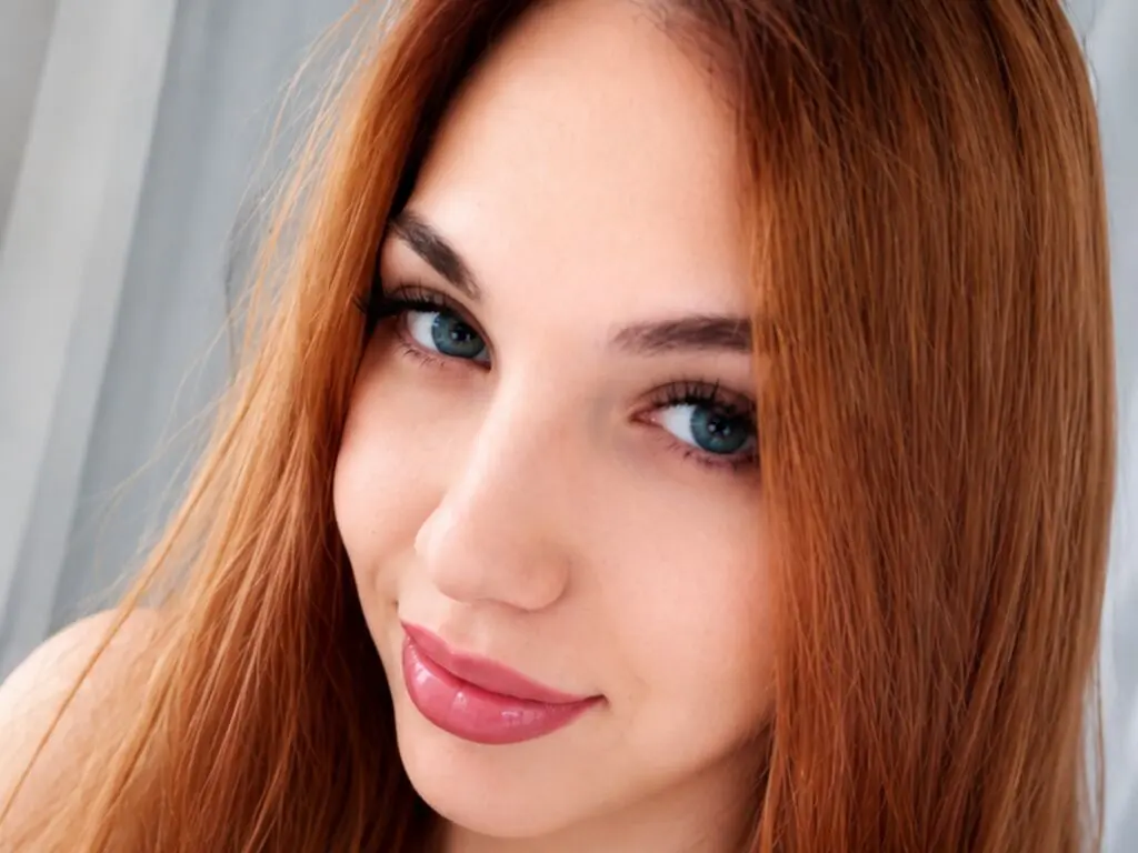 Close-up portrait of a smiling young woman with long red hair and blue eyes in a bright indoor setting.