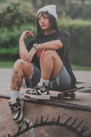 Trendy young woman in black t-shirt and denim shorts sitting on a skateboard at a skatepark with a cool aesthetic.