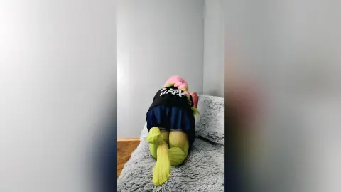 EstherTrix in a black top and blue skirt, kneeling on a grey shag rug with her head down.