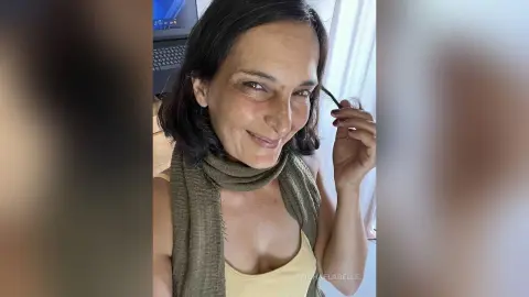 MichaelaBelle smiles while wearing a yellow tank top and an olive green scarf, tucking a strand of dark hair behind her ear.
