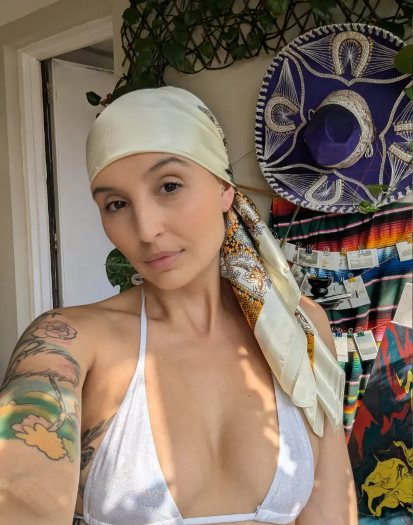oh_charlie poses with a white headwrap, showing her bare shoulders and chest in a white top against a colorful patterned background.