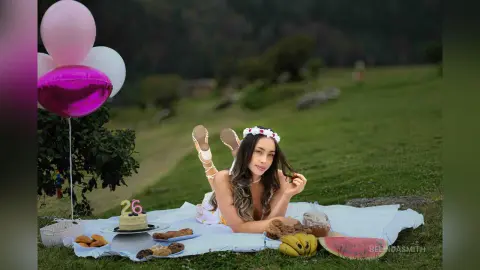 Young woman in a white floral dress relaxing on a picnic blanket with pink balloons in a lush green meadow.
