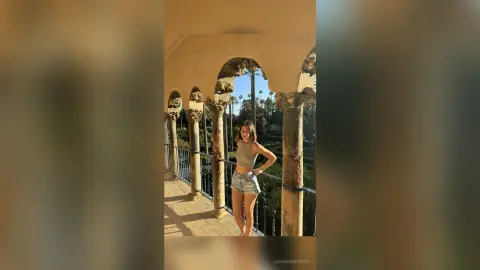 LuanaRodgers poses in a grey tank top and denim shorts on a sunny balcony with stone arches.