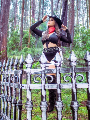 Cowgirl in black fringe outfit and hat posing by a white picket fence in a lush green forest.