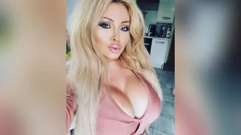 Glamorous blonde woman in a pink v-neck top posing in a bright modern kitchen.