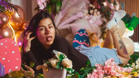 Young woman in a black top and denim shorts relaxing amidst pink balloons and flowers in a festive setting.