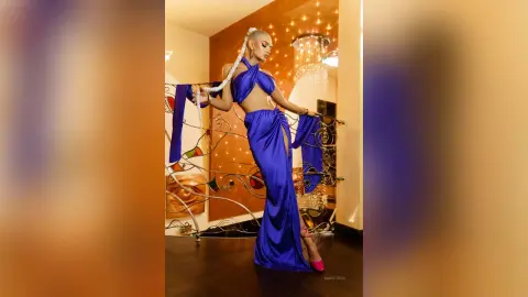 Anastasia poses in a blue halter-neck maxi dress against a decorative metal railing and warm lighting.