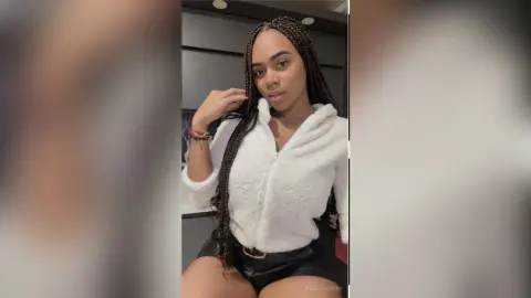 PamelaCreed wears a white zip-up fleece jacket and black leather shorts, posing with her long dark braids draped over her shoulder.