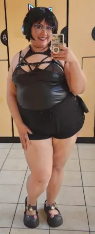 Curvy woman in black cosplay outfit with cat ears posing in a locker room setting.