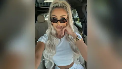 AlexaEvans poses in a car wearing sunglasses and a white t-shirt, showing off her long blonde wavy hair and gold watch.