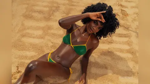 Smiling Black woman in a green crochet bikini relaxing on a sunny sandy beach.