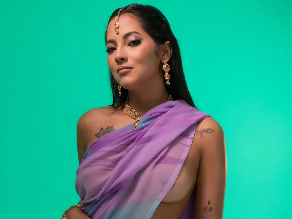 Beautiful woman in purple traditional Indian saree and gold jewelry posing against a teal background.
