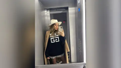 VictoriaWynn poses for a mirror selfie in an elevator, wearing a white hat, a black sweatshirt with a white logo, and a tan coat.