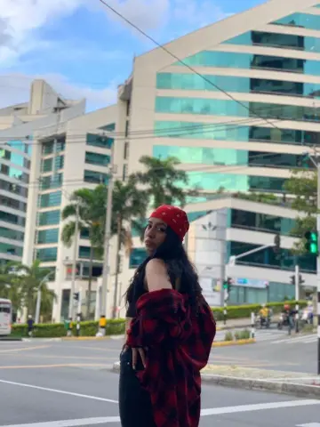 Young woman in a red bandana and black outfit standing on a city street by modern glass buildings.
