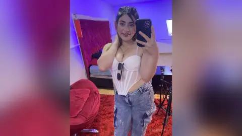 Young woman in a white corset top and denim jeans taking a selfie in a moody, purple-lit bedroom.