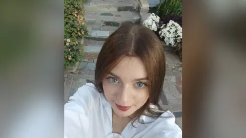 Smiling young woman in a white shirt posing outdoors on stone steps near garden flowers.
