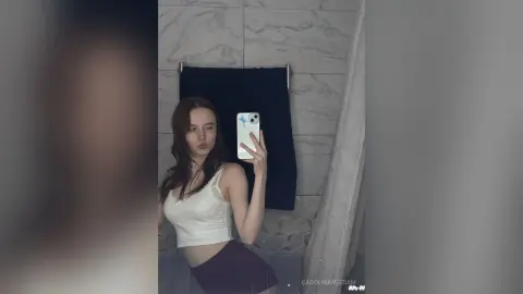 Young woman in a white ribbed tank top taking a mirror selfie in a modern marble bathroom.