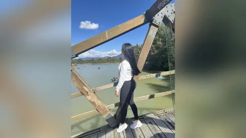 Woman in white long-sleeve shirt and black leggings standing on a wooden bridge overlooking a scenic lake.