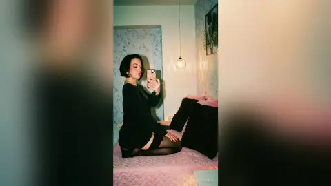 Young woman in a black long-sleeve top taking a mirror selfie in a cozy, dimly lit bedroom.