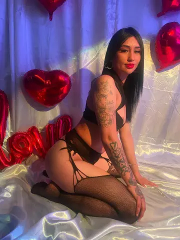 Tattooed woman in black lingerie and fishnet tights sitting among red heart balloons in a romantic setting.