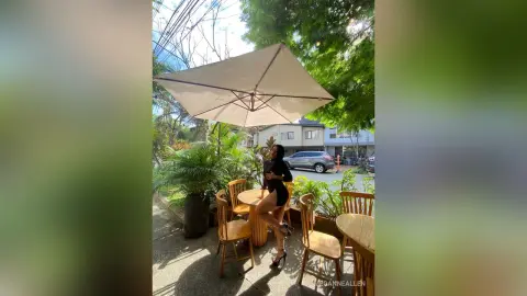 MeganneAllen poses outdoors in a black bodysuit, sitting on a wooden chair at a patio table under a large white umbrella.