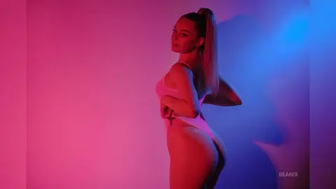 BeaNix poses in a pink bodysuit against a pink and blue lit background, looking back over her shoulder.