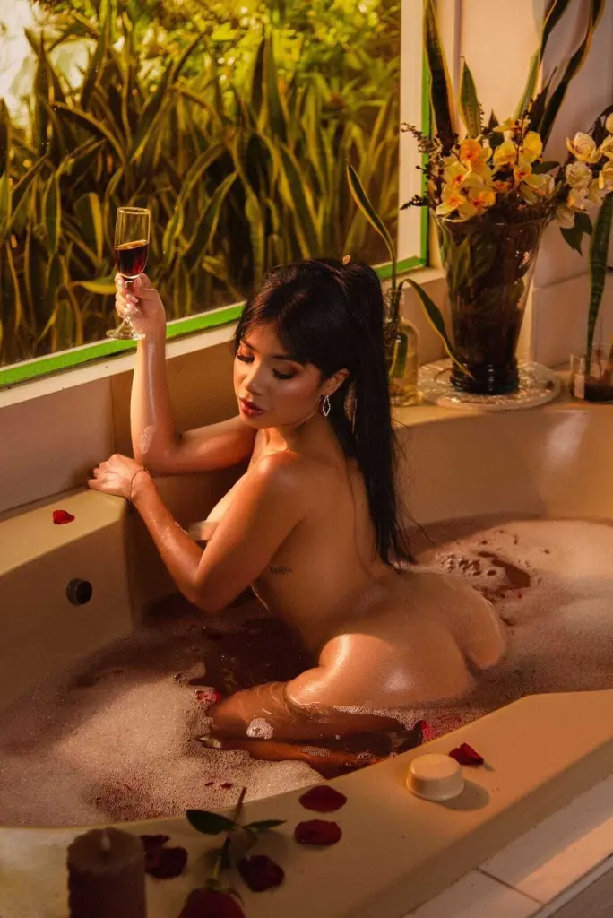 ArianaWoods poses in a white bodysuit against red curtains, smiling while sitting near lit candles.